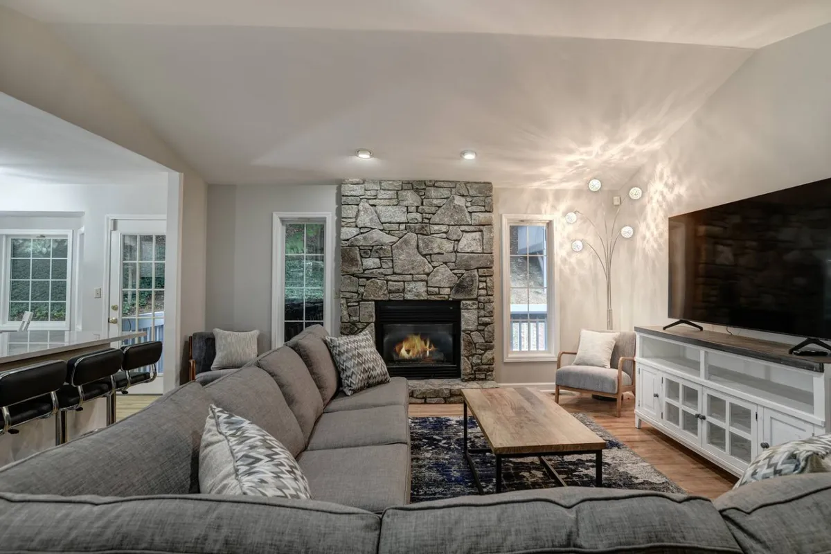 The image centers on a stone fireplace surrounded by a large sectional sofa, coffee table, TV, and decorative lighting. The atmosphere is warm and inviting, perfect for group relaxation and evening gatherings.