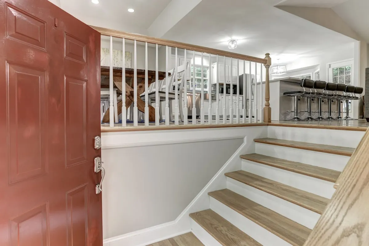 This image features the home's inviting red front door, a staircase, bar stools at a nearby counter, and a glimpse of the dining area. Sunlight streams through the windows, creating a welcoming transition between inside and out.