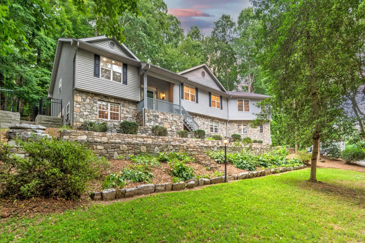 The image features the stately stone facade of a single-family house set amidst a beautifully landscaped garden in Biltmore Forest, North Carolina. A spacious front porch invites guests to relax and enjoy the serene surroundings, while lush greenery and manicured flowerbeds frame the scene, evoking a sense of peaceful retreat and inviting outdoor gatherings.