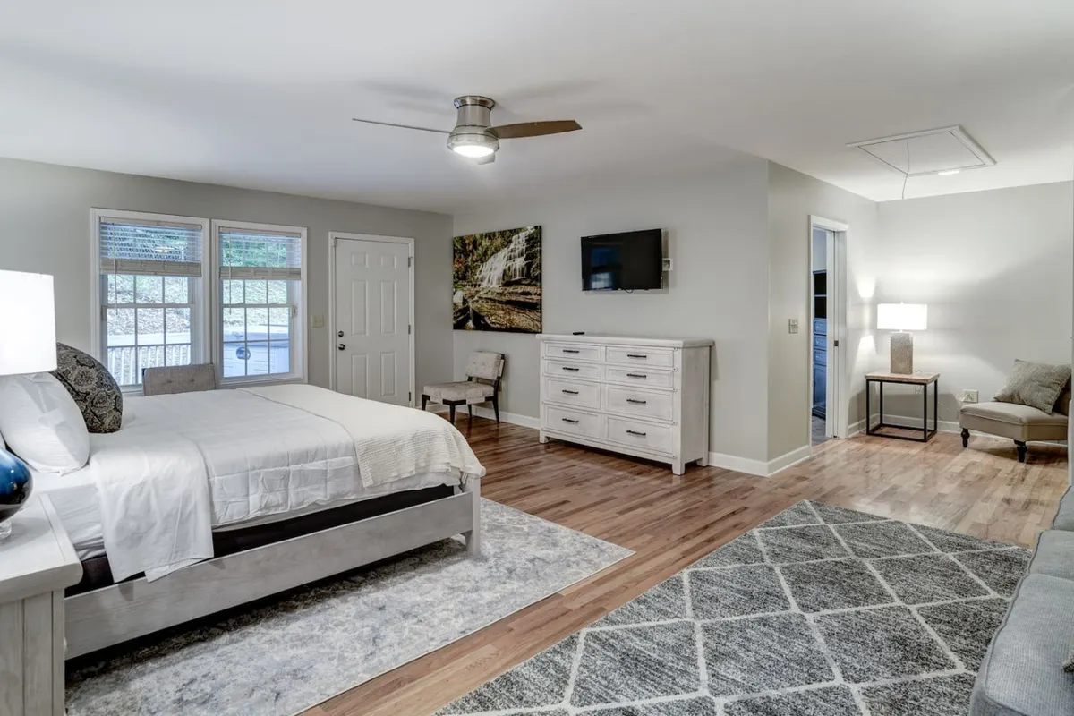 A serene bedroom featuring a plush bed with a textured rug underneath, large windows letting in soft natural light, a dresser, stylish artwork, and a cozy sitting area. The space feels open and inviting, perfect for unwinding after a long day.