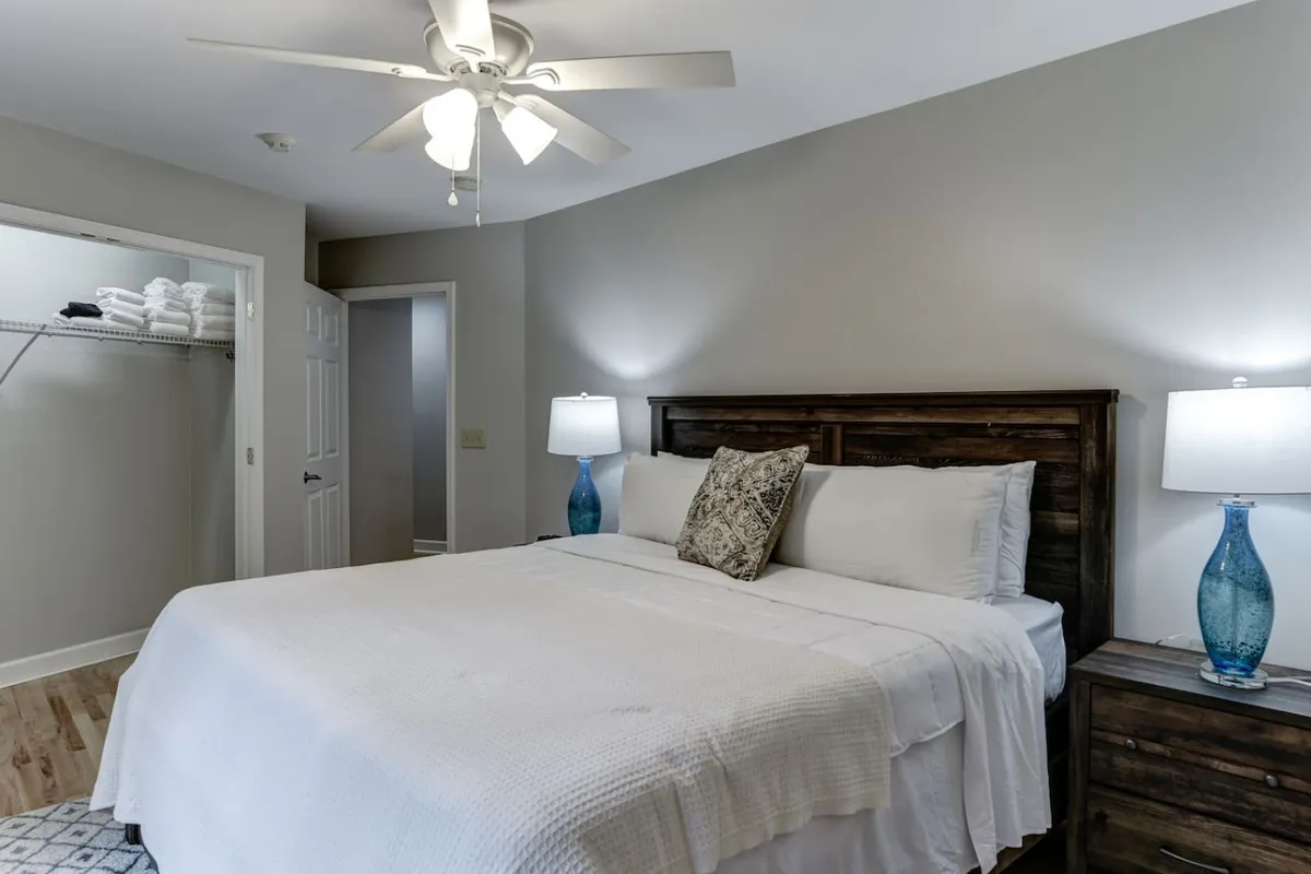 A tranquil bedroom centered on a comfortable bed adorned with decorative pillows, flanked by two bedside lamps. An open closet with neatly folded towels adds a sense of convenience and readiness.