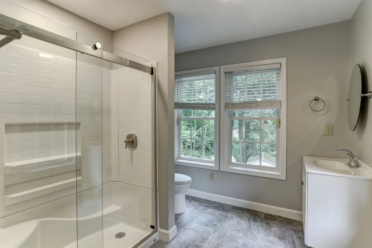 A modern, light-filled bathroom featuring a glass-door shower, a sleek vanity with sink, and large windows. The space is airy and calming, ideal for refreshing morning routines or unwinding at night.