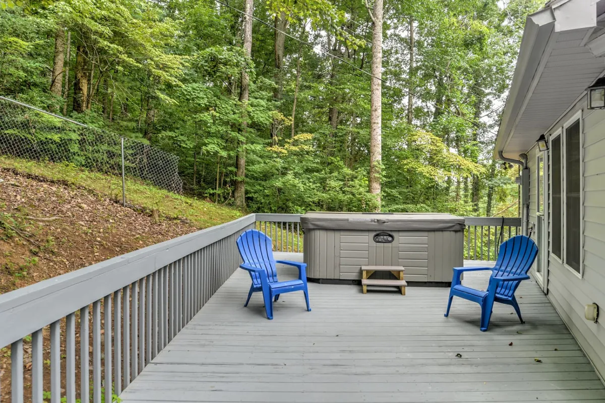 A peaceful exterior view of a private hot tub surrounded by lush greenery, with blue chairs nearby inviting guests to relax. The deck setting is enveloped by nature, creating a tranquil retreat for soaking under the stars or enjoying a quiet moment outdoors.
