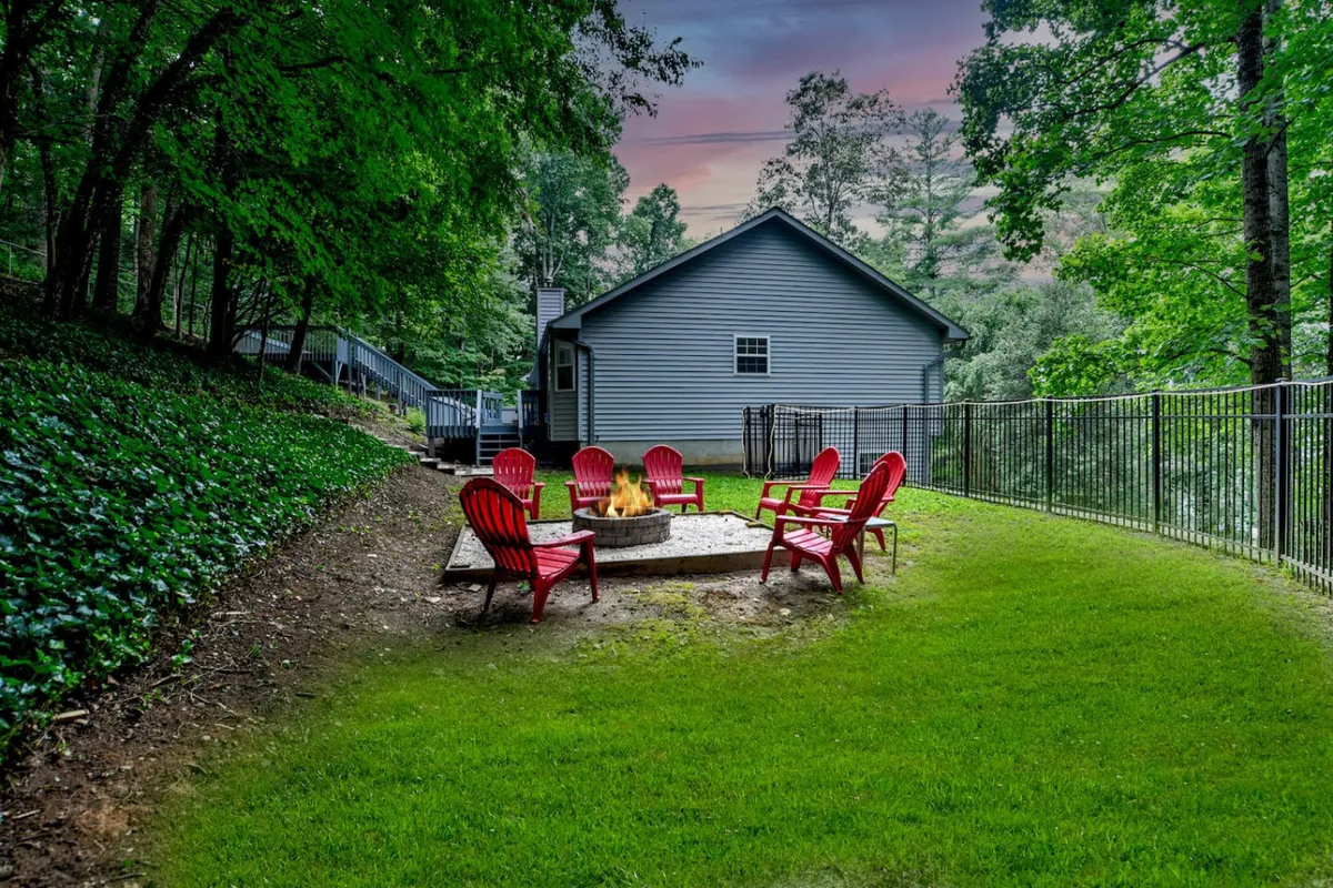 A cozy fire pit area featuring red chairs arranged in a circle on a green lawn, bordered by tall, leafy trees. The setting is ideal for evening gatherings, storytelling, and sharing s’mores under the open sky.